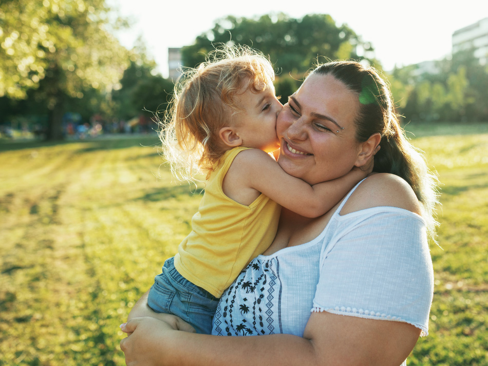 Child kisses smiling woman in sunny park.