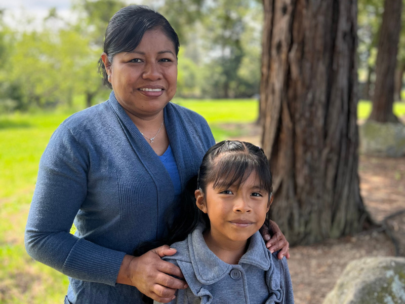 Mother and daughter smiling outdoors near tree.