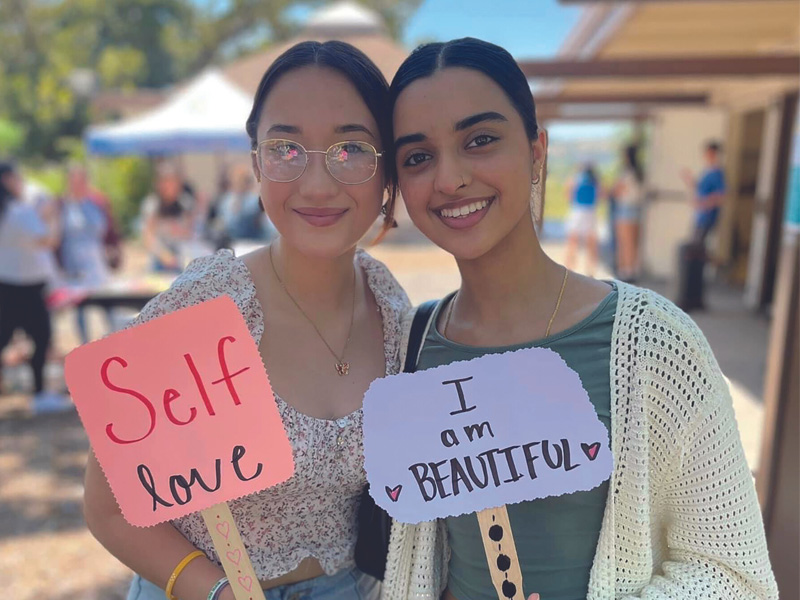 Two women holding self-love signs, smiling outdoors.