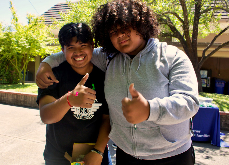 Two friends smiling with thumbs up outdoors.