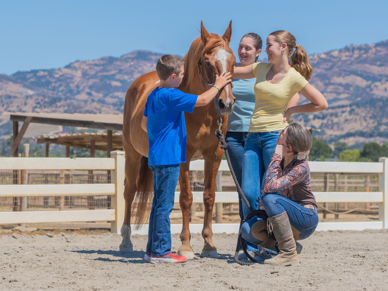 Family petting horse at ranch with mountain backdrop.