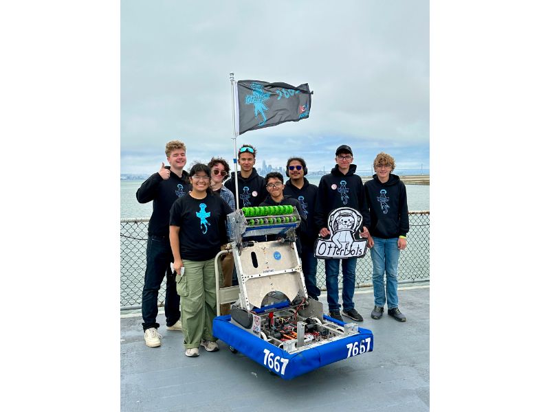 Team posing with robot and flag by water.